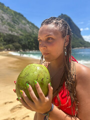 Young woman in a red bikini with braided hair drinking from a fresh coconut on a sunny tropical beach, relaxed and cheerful in warm natural light.