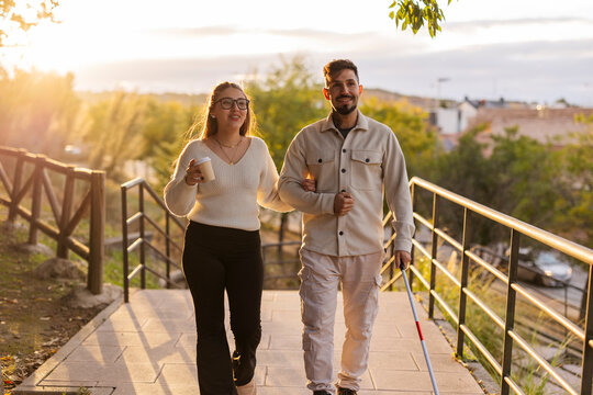 Woman guiding blind man walking with white cane at sunset