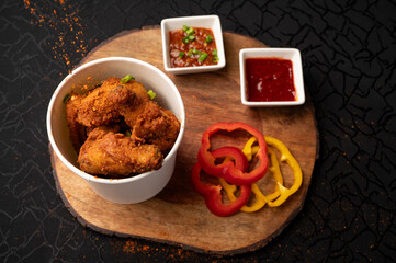 Top view of chicken wings on a wooden board as masala is sprinkled over them, served with red chilli sauce and a creamy dip, creating a bold and flavorful food presentation on a dark black background.
