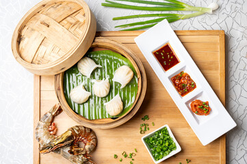 Top view of prawn hargao dim sum arranged in a bamboo steamer on a wooden board, styled with cut green chillies, red chilli sauce, assorted dipping sauces, and raw prawns for a fresh presentation