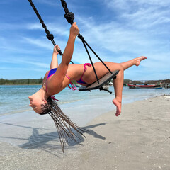 A joyful young woman with braided hair enjoys a tropical beach swing, leaning back with a bright smile over clear turquoise water and light sand in sunny weather.