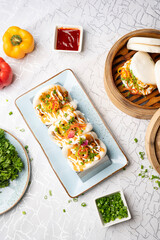 Top view of pulled butter chicken bao with pickled onions and tandoori mayo paired with crispy chilli chicken bao and hot garlic sauce, styled with red chilli sauce, cut green chillies, capsicums