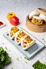 Top view of pulled butter chicken bao with pickled onions and tandoori mayo paired with crispy chilli chicken bao and hot garlic sauce, styled with red chilli sauce, cut green chillies, capsicums