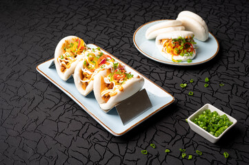 Pulled butter chicken bao with pickled onions and tandoori mayo served in a tray alongside crispy chilli chicken bao with hot garlic sauce in a plate, styled with cut green chillies in a small bowl