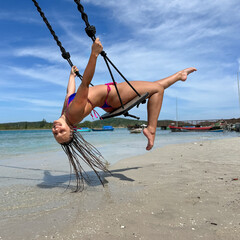 A joyful young woman with braided hair enjoys a tropical beach swing, leaning back with a bright smile over clear turquoise water and light sand in sunny weather.