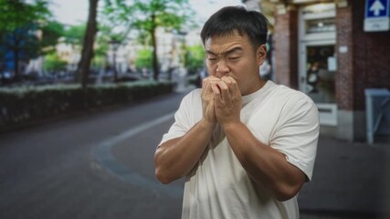 Man rubbing hands while looking anxious on city street in front of brick storefront with blurred pedestrians and parked bicycle; anxiety. - Powered by Adobe