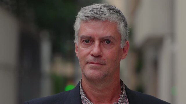 Portrait of middle-aged man with gray hair and neutral expression outdoors, showing confidence and realism with natural light and background blur