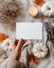 Cozy Autumn card mockup. Flat lay still-life with white pumpkin, dried leaves, lavender and lit candle. Blank card held in hand. For personalized greeting card, invitation or seasonal promotion.