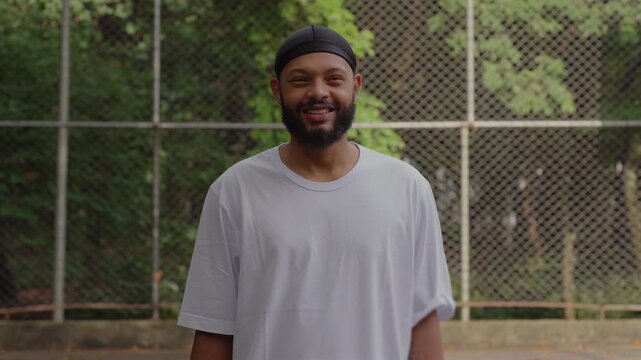 Smiling African American man wearing durag standing outdoors in basketball court, looking at camera with joyful expression and relaxed confident body language