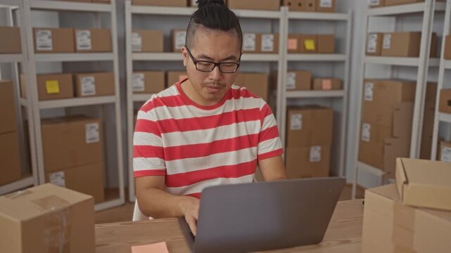 Man typing on laptop at a wooden table surrounded by cardboard parcels and shelving in a building; concentration.
