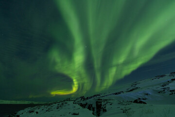 Spectacular wide arc of intense green Aurora Borealis soaring high above a snow-covered rocky ridge...
