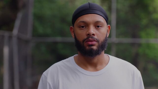 Bearded African American man wearing durag standing outdoors, looking directly at camera with serious composed expression against green blurred background in urban park setting