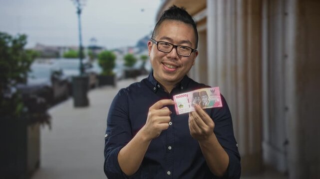 Man holding indonesian rupiah banknote between fingers on city street, smiling and pointing with hand near chest; confidence prosperity.