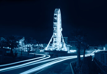 Long exposure light trails of car headlights on a winding road at night - White ferris wheel at an...