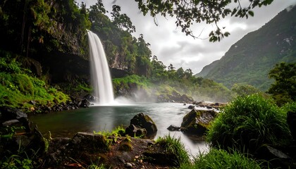 Waterfall cascades into a calm pool surrounded by lush greenery and volcanic rocks on a cloudy, overcast day