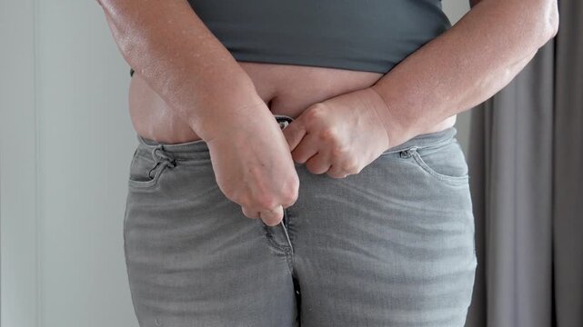 Fat woman adjusting jeans during morning routine with visible frustration. Woman struggling with jeans, attempting to fasten tight button over exposed fat stomach, hands pressing