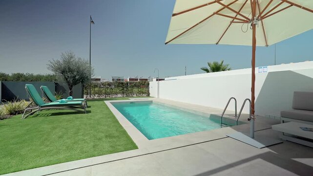 White parasol shading a comfortable outdoor sofa and sunbeds next to a turquoise private swimming pool in a contemporary house with artificial grass