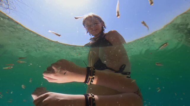 Fascinating underwater split shot of smiling young woman in bikini enjoy natural pedicure and skin cleaning from school of small garra rufa fish in clear turquoise water