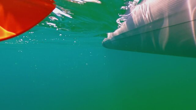 Unique split-screen underwater perspective showing red paddle propelling grey kayak through tranquil, clear green sea, with bubbles rising to shimmering surface