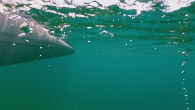 Split-screen underwater view of paddleboard and an orange paddle moving through clear turquoise water, with bubbles forming on surface as board glides forward on sunny day