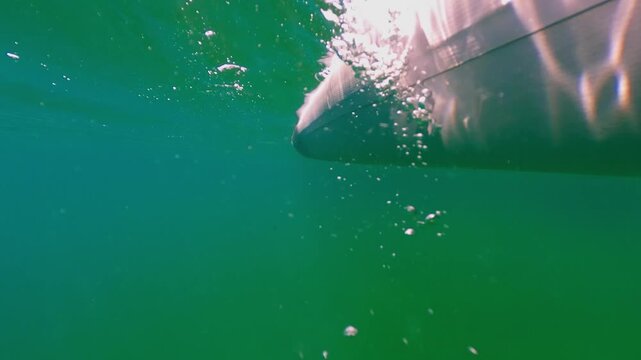 Underwater perspective of an orange kayak paddle rowing next to boat hull, creating bubbles and movement through clear green water of lake or sea on sunny summer day