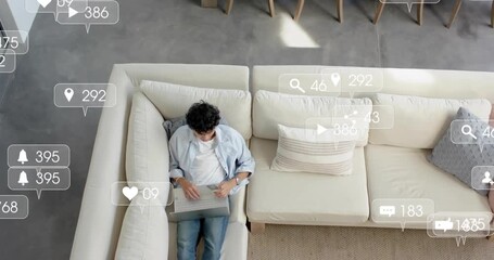 Man sitting on sofa using laptop, posting and receiving social media notifications flooding room - Powered by Adobe