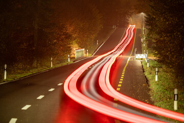 Lonely road in the evening. Cars are driving on the road through the forest. Tail lights are producing red light trails. Reflections on the wet road.