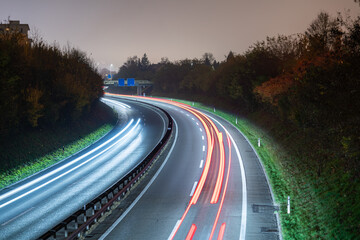 Highway at night, under a grey foggy sky, with light trails of passing cars. Green meadow and trees on the sides.