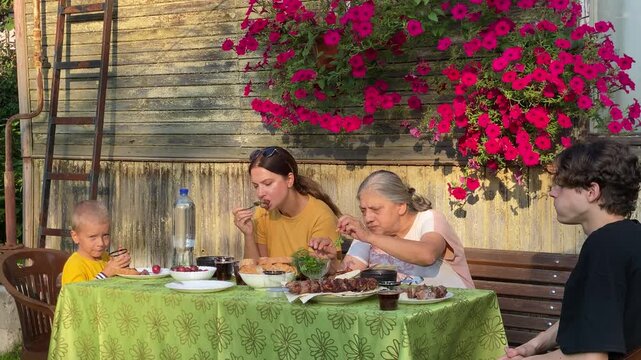 A three-generation family&mdash;grandmother, mother, and two children&mdash;sits around an outdoor table, enjoying a delicious barbecue meal. This summer dinner is taking place outside an old, rustic wooden house