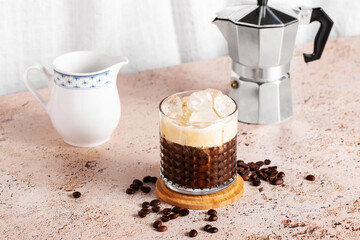 Iced creamy coffee into a glass with moka pot and milk jug on beige kitchen table background. Coffee break during a hot summer day, refreshing drink