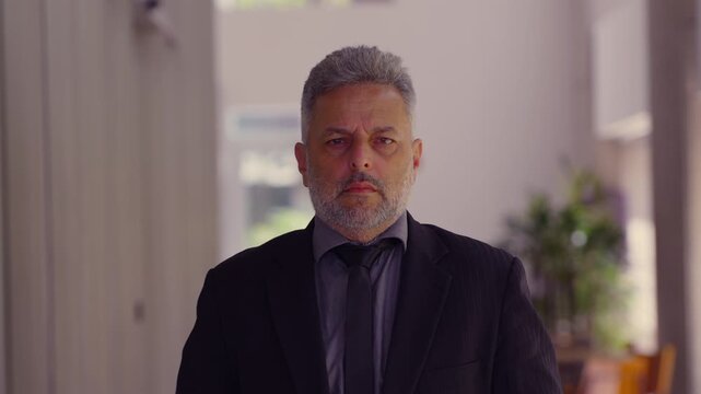 Portrait of middle-aged man in suit standing indoors with serious and determined expression, confident posture suggesting authority and introspection