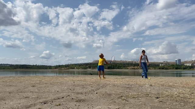 A young boy and a woman are playing badminton on the dry, dusty shore of a lake under a wide sky with scattered clouds. The boy bends down holding a racket, while the woman looks on.
