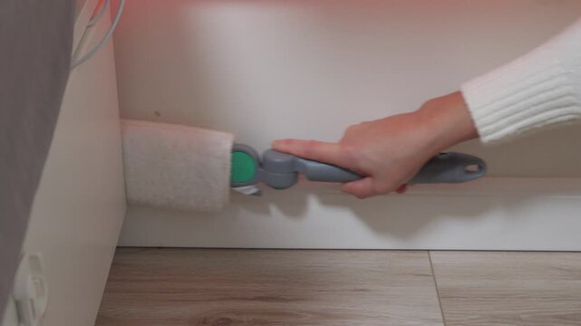 A hand uses a handheld roller-style cleaning tool to scrub the wall or baseboard in the small, tight gap behind furniture. This demonstrates a dedicated effort for thorough home cleaning.