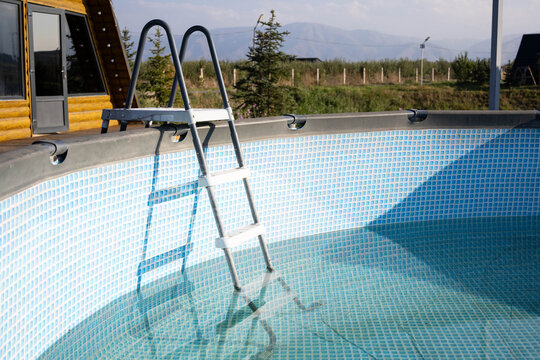 A metal pool ladder stands inside a partially filled outdoor above-ground swimming pool with clear water and a mountain landscape in the background