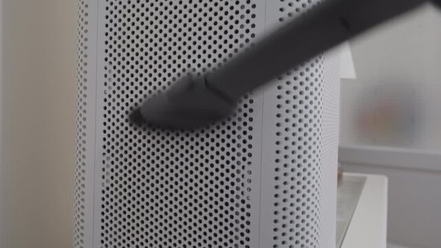 A handheld vacuum brush cleans perforated outer casing of a white, cylindrical air purifier. This routine maintenance is essential for keeping the appliance running efficiently and ensuring clean air.