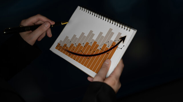 Hands holding a notepad featuring a bar graph and a rising arrow that symbolize growth and progress in business or financial analysis against a dark background. Scalp - Powered by Adobe