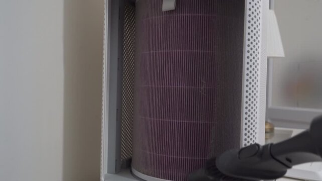 A handheld vacuum cleaner attachment removes heavy dust from the pleated, purple filter of a home air purifier. This maintenance ensures clean indoor air quality and device efficiency.