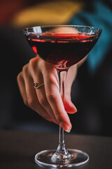 Elegant, deep red cocktail held in a coupe glass with a feminine hand and silver ring, dark background, shallow depth of field, moody atmosphere