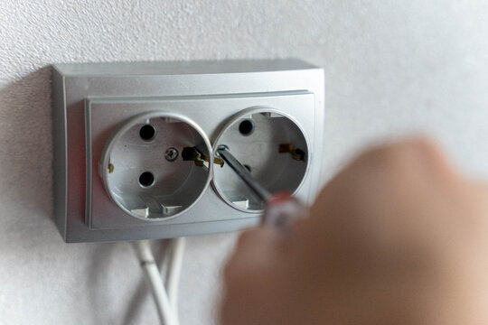Close-up of a hand using a screwdriver to repair an electrical outlet with dual sockets on a textured wall, showcasing home maintenance and DIY skills
