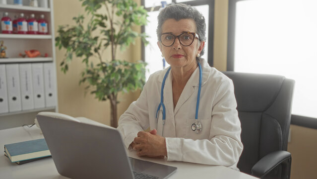 Senior woman doctor with grey hair sitting seriously with crossed arms in modern clinic office using laptop and wearing stethoscope around her neck.