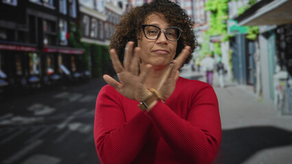 Fototapeta premium Woman gently touches curly hair in red sweater on a bright sunny bustling city street; quiet confidence.