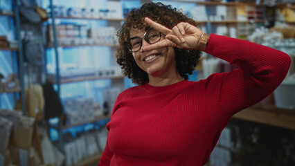 Woman hand making peace sign in a colorful home decor shop interior with shelves of pottery, candles, and textiles displayed around her; joy.