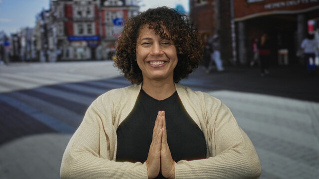 Middle aged hispanic woman smiling and pressing hands together in prayer gesture on a busy city street; serenity reflection.
