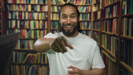 Man in white t shirt shielding eyes with hand while smiling and gesturing toward bookshelves in a building library aisle; surprise discovery learning.