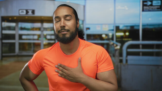 Man with hand on chest exhaling at airport terminal outdoors in bright orange tshirt, eyes closed and one hand on hip; relief recovery.