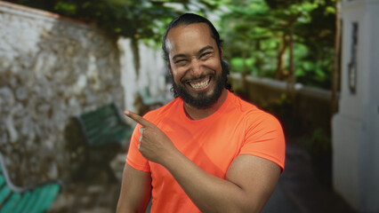 Fototapeta premium Man points finger to the side on a street near a stone wall and bench while wearing a bright orange shirt and ponytail; playful surprise.
