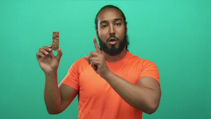 Man in bright orange shirt holding granola bar and points finger to snack in green studio; health...