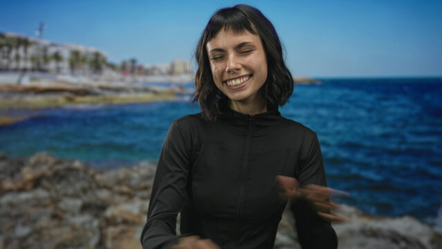 Athletic woman in black jacket smiles and crosses arms at rocky seaside coast under clear blue sky; joy.