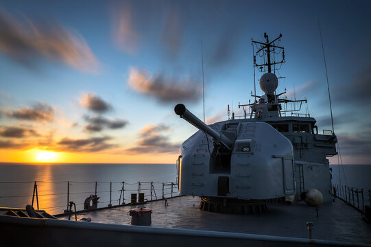 A warship sails in a calm sea at sunset.
