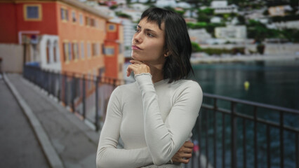 Young hispanic woman with hand on chin gesture on a colorful street lined with buildings; quiet reflection.